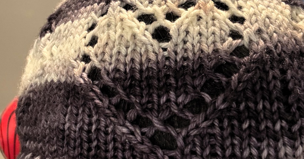 A red knitted beanie with a folded brim and lace heart motif shown outdoors.